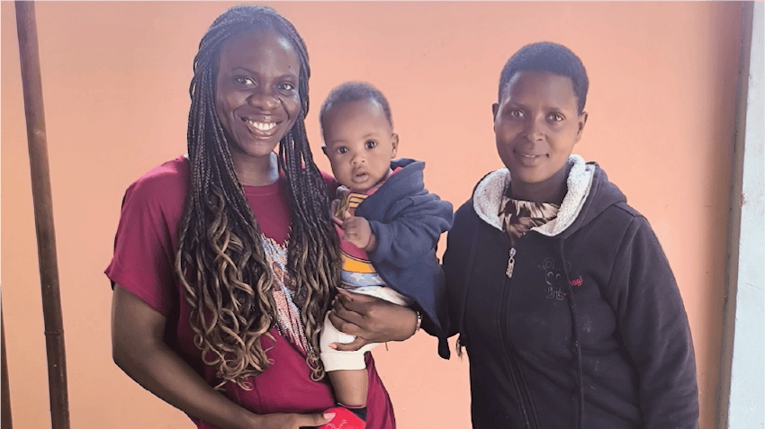 Glodie, an intern in Arusha, Tanzania, smiles and stands next to a young mother, she is posing and holding the mother's child