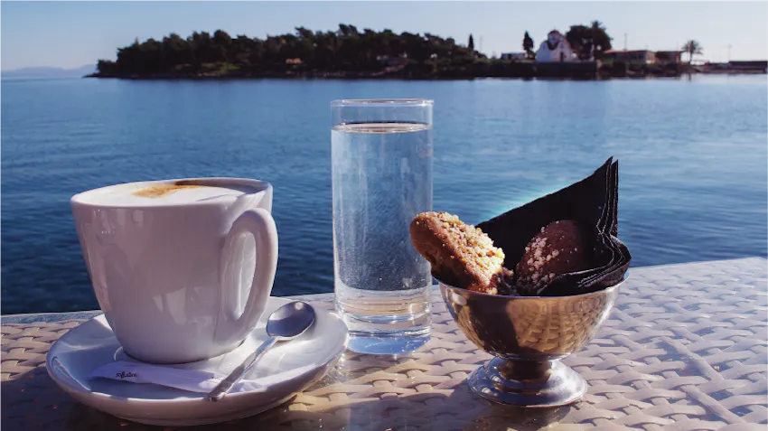 A cup of Greek coffee, a glass of water, and a sweet pastry treat are photographed, on a table, in front of the blue water and sky.g