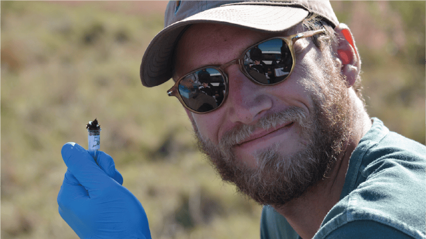 Nick smiles at the camera and holds up a sample, taken from his environmental conservation work in South Africa