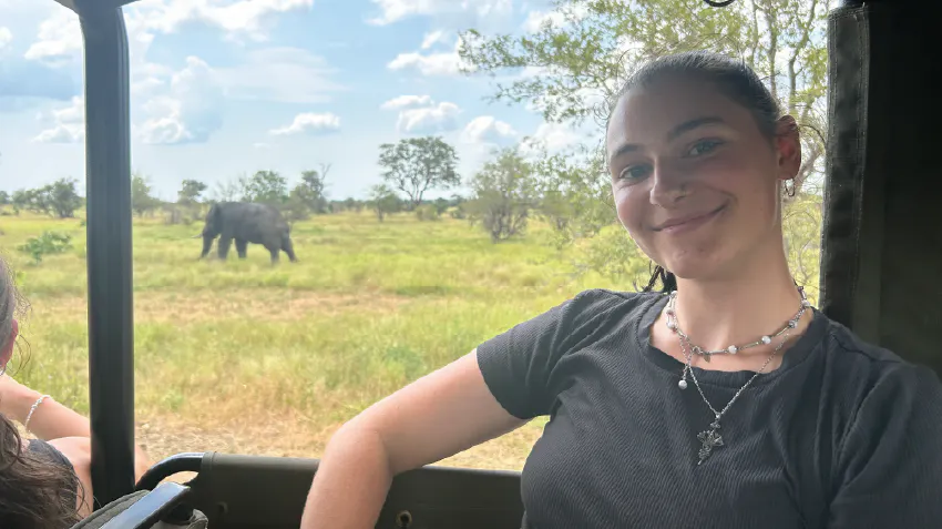 Emily is pictured posing in the window of a vehicle, with an elephant pictured in the distance behind her