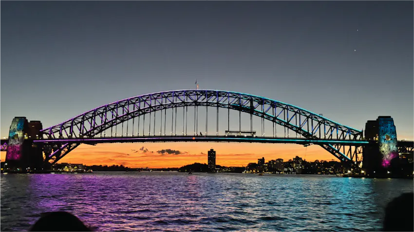A photo of the Sydney Harbour Bridge, taken in the evening and lit up by brilliant sunset colours over the sparkling water