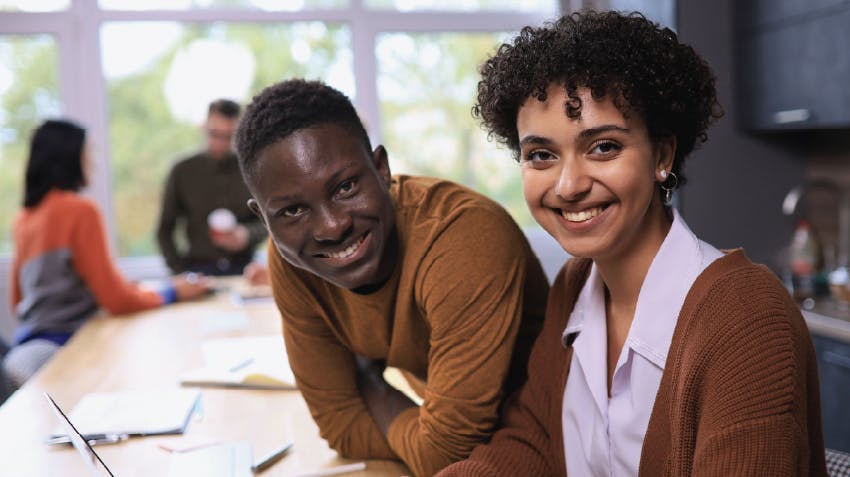 Two students posing together at a desk, within a classroom, looking toward the camera and smiling