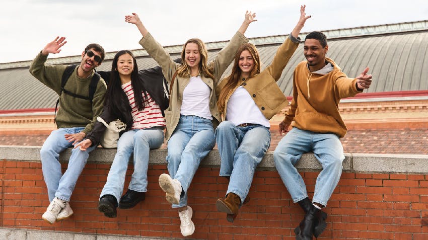 A group of students sitting together on a brick wall and smiling, with raised arms