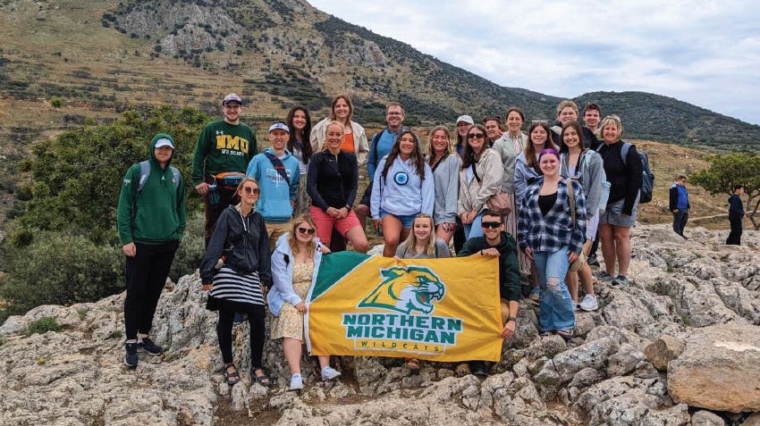 A group of students from NMU pose for a photo in Greece, during an Intern Abroad HQ study abroad program; holding a flag with the text, Northern Michigan Wildcats