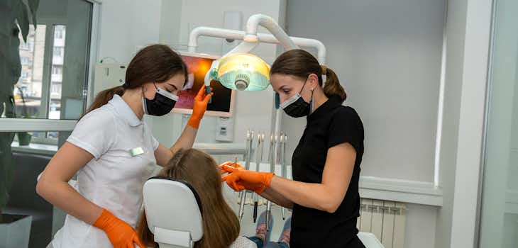 A dentist works with a patient, accompanied by a dentistry assistant.