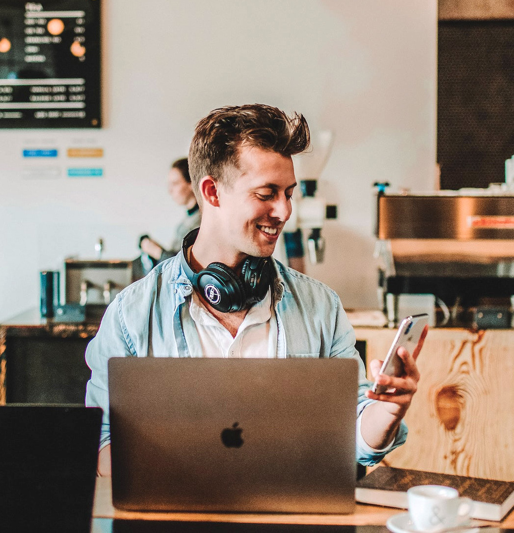Remote intern works from a cafe, smiling while checking his phone with a laptop open.